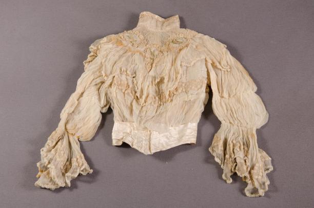     Blouse van witte gaze de soie gegarneerd met creme passement met gekleurde medaillons en wit satijnen ceintuur., collectie Centraal Museum