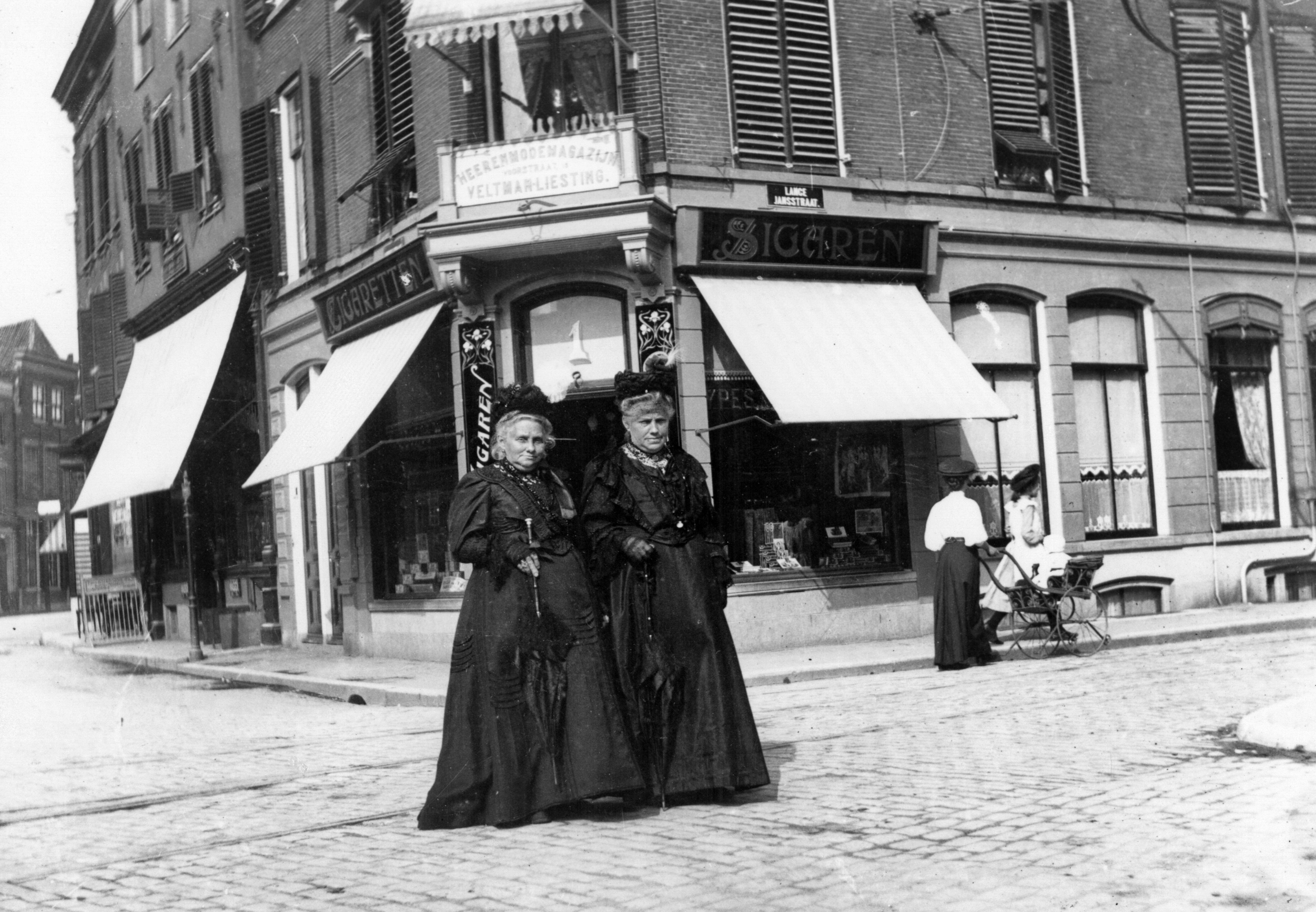 De kanten details op de jurken, de hoedjes met voiles en de parasols met chique handvatten maken duidelijk dat deze dames uit de welgestelde klasse komen. De winkel waar zij voor staan ‘Heerenmodemagazijn Veltman Liesting’ bestaat anno 2025 nog steeds. (Foto Joh. A. Moesman, nr. 72580)