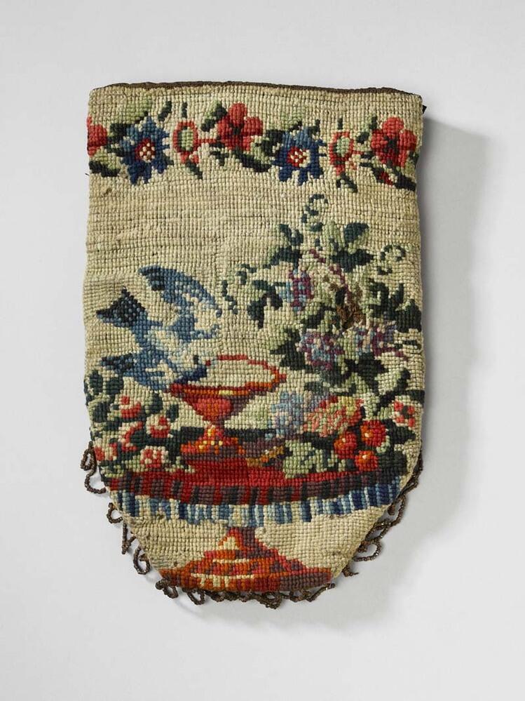 Tasje in tapisserie waarop afgebeeld een tafel met vaas waaruit vogel drinkt, bloemstuk en bloemrand