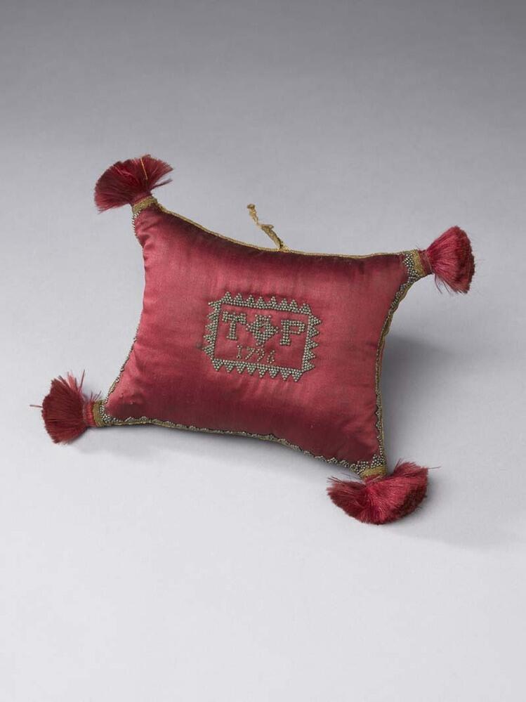 Een speldenkussen van rood satijn met aan de zijkanten afgezet met goudband, in de hoeken kwastjes
