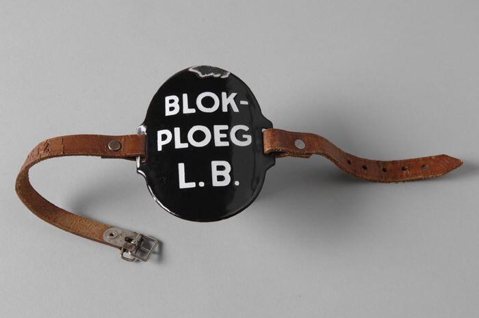 Ovaal zwart geëmailleerd armschildje met "BLOK-PLOEG L.B.", met leren riempjes aan weerszijde