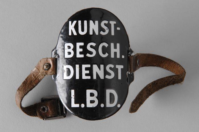 Ovaal zwart geëmailleerd armschildje met "KUNST- BESCH. DIENST L.B.D.", met leren riempjes aan…