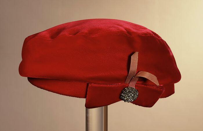 Ronde baret, rood fluweel met voor platte strik met roze ripslint waarop rozet van metalen kraaltjes