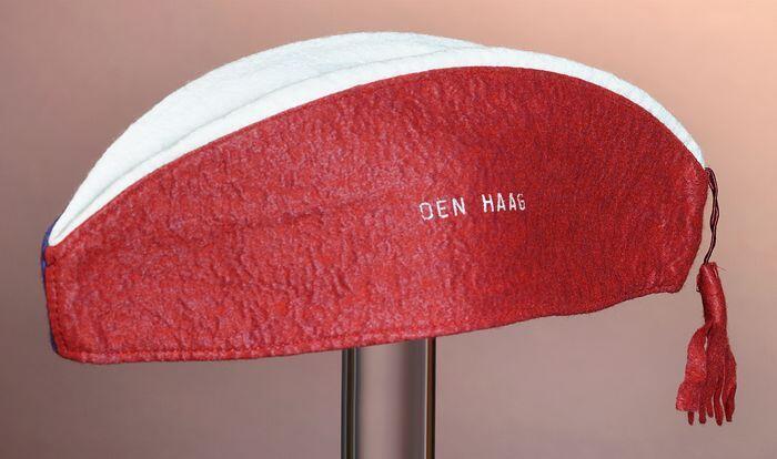 Ovale vakantiemuts (?) van blauw, rood en wit vilt met rood kwastje, rood zijvlak met "DEN HAAG"