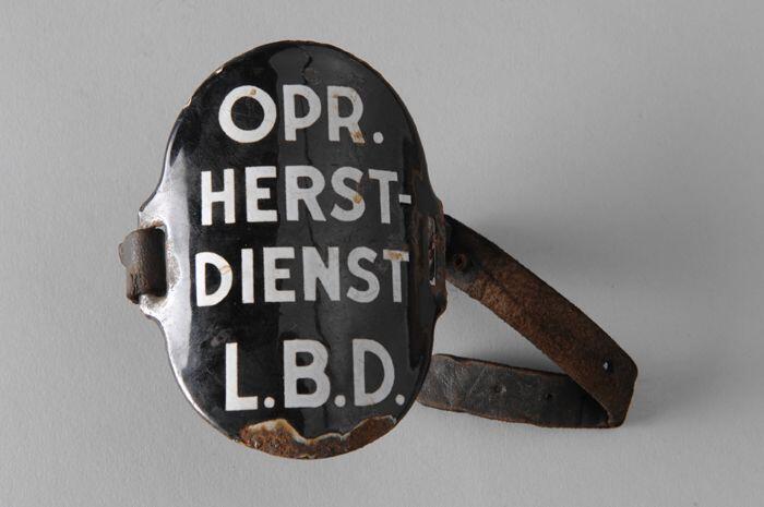 Ovaal zwart geëmailleerd armschildje met "OPR. HERST-DIENST L.B.D.", met (gebroken) leren riempjes…