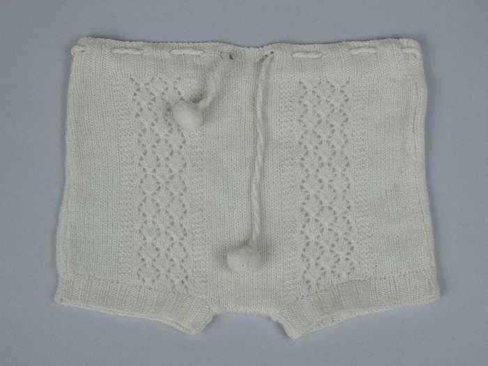 Korte babybroek gebreid van crèmekleurige dunne wol in tricotsteken en rechte banen ajour, koordje…