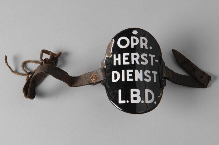Ovaal zwart geëmailleerd armschildje met "OPR. HERST-DIENST L.B.D.", met leren riempjes aan…