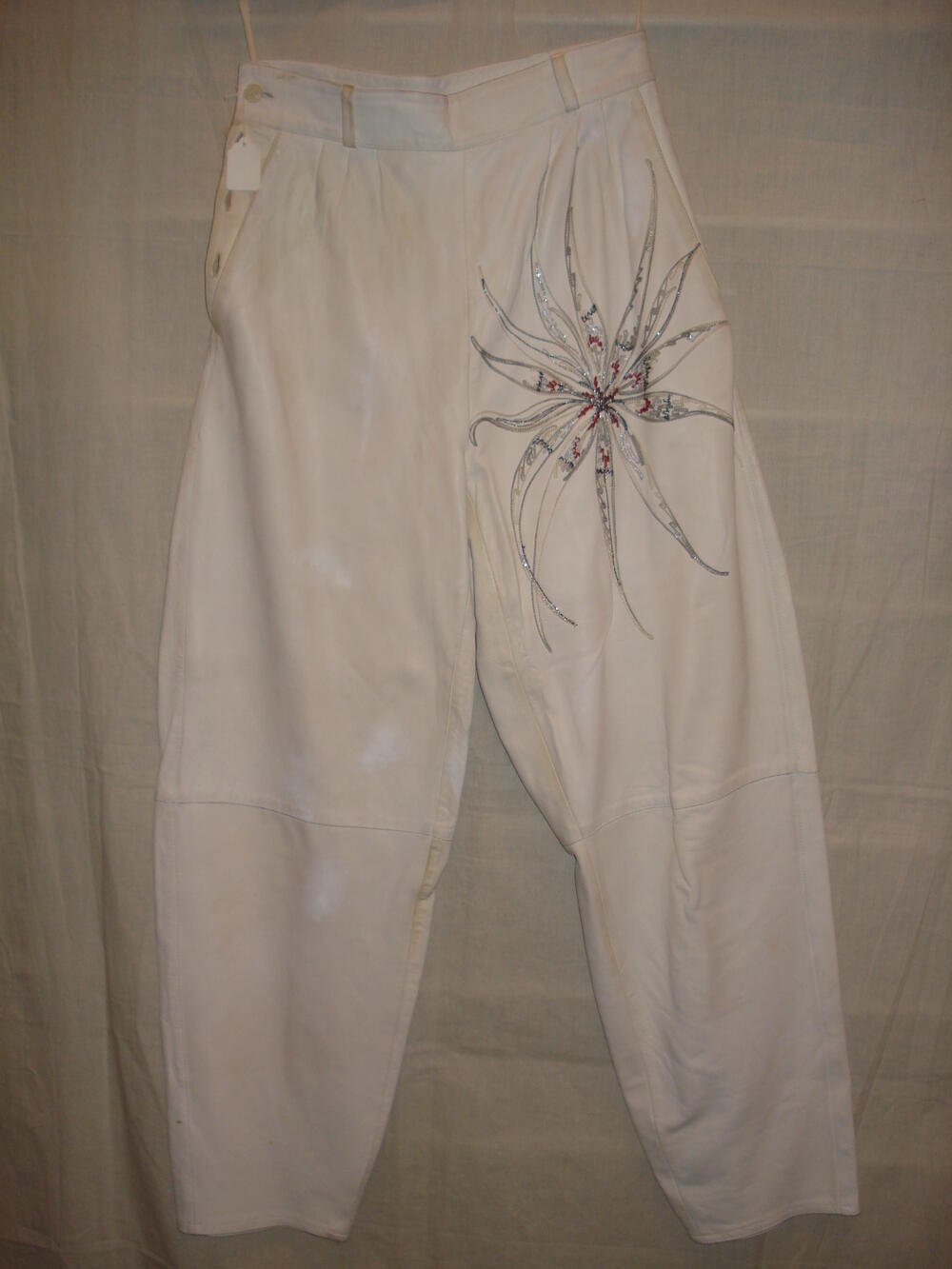 Broek van wit leer met op het linker bovenbeen een geborduurde bloem met zilver en rood,  onderdeel…