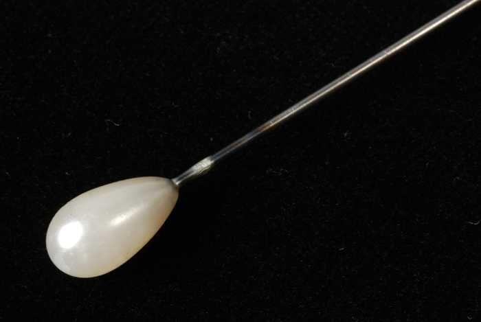 Hoedenspeld, korte metalen pen met scherpe punt, druppelvormige witte sierknop met parelmoerglans