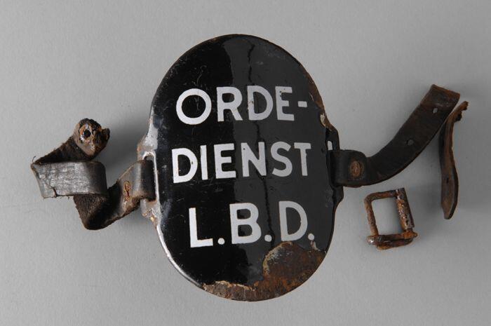 Ovaal zwart geëmailleerd armschildje met "ORDE-DIENST L.B.D.", met leren riempjes aan weerszijde