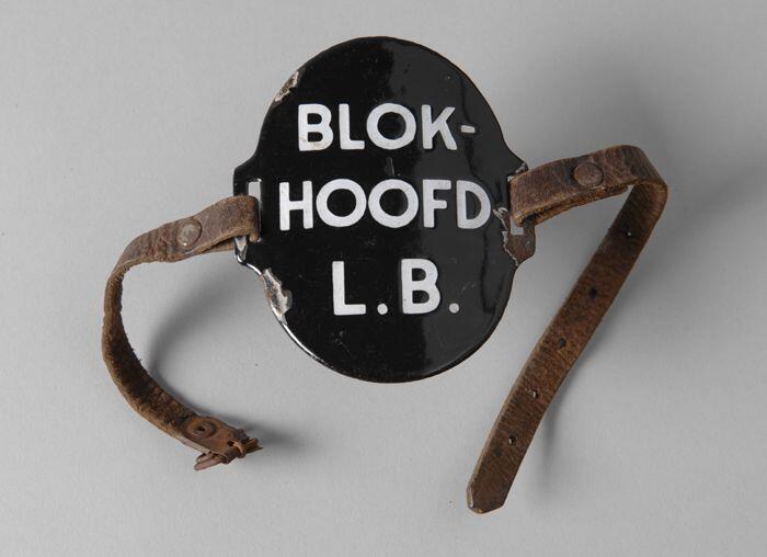 Ovaal zwart geëmailleerd armschildje met "BLOK-HOOFD L.B.", met leren riempjes aan weerszijde
