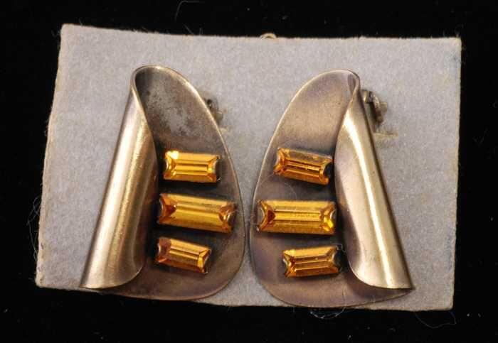 Paar oorbellen, clip met omgebogen metalen blad waarop drie oranje glastaafjes, op grijs…