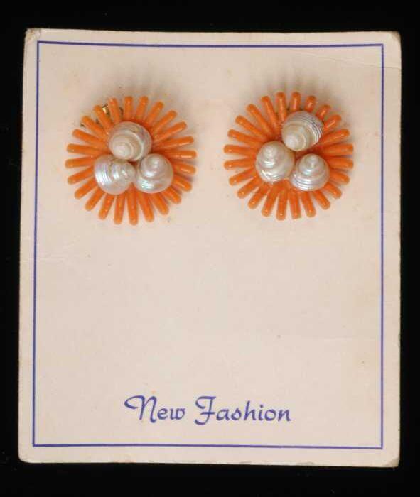 Paar oorbellen, clips met oranje plastic straalvormige bloem met een hart van schelpjes, op karton,…