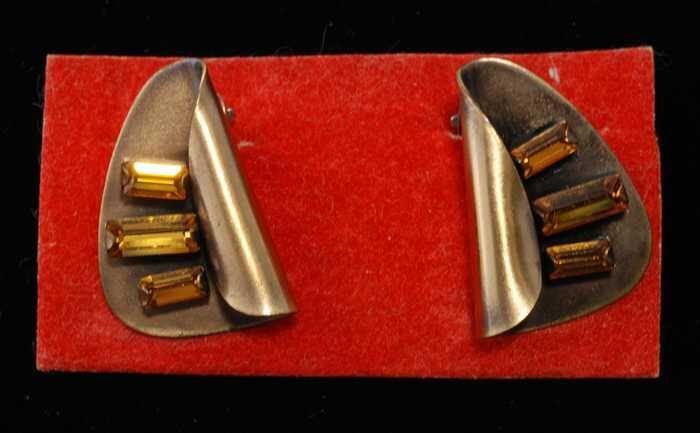Paar oorbellen, clip met omgebogen metalen blad waarop drie oranje glastaafjes, op rood…
