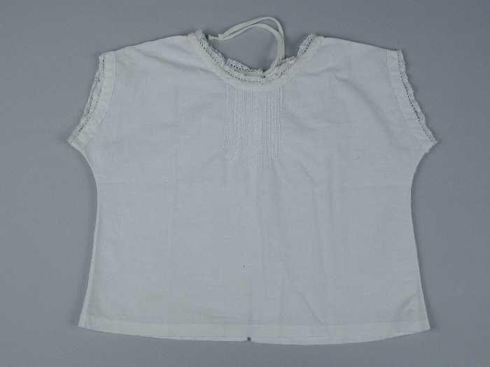 Babyhemd van wit katoen ronde hals, korte aangeknipte mouw, achter open, smalle rand kant langs…