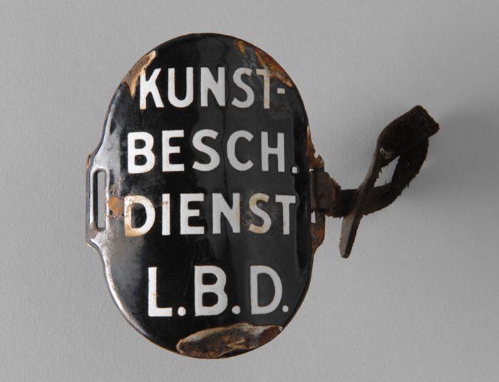 Ovaal zwart geëmailleerd armschildje met "KUNST- BESCH. DIENST L.B.D.", met één leren riempje
