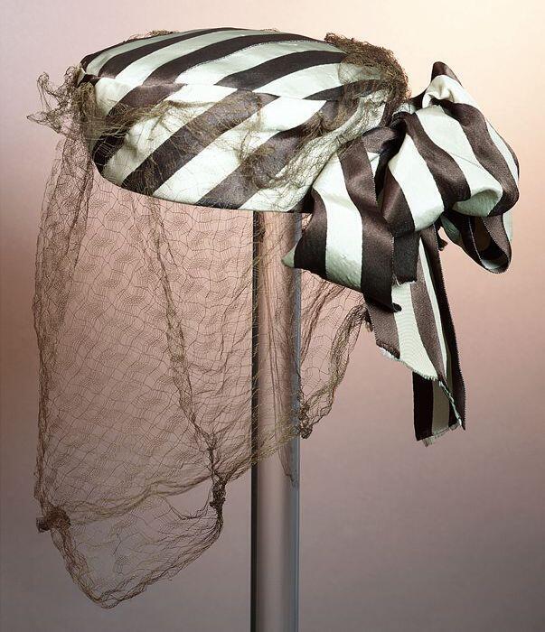 Ronde dameshoed van bruin-beige gestreepte glanzende stof met grote strik achter en bruine voile