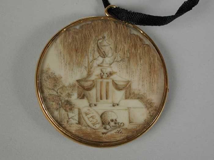 Memoriestukje van haarin medaillon van glas gevat in gouden lijst, hangend aan smal zwart zijde…