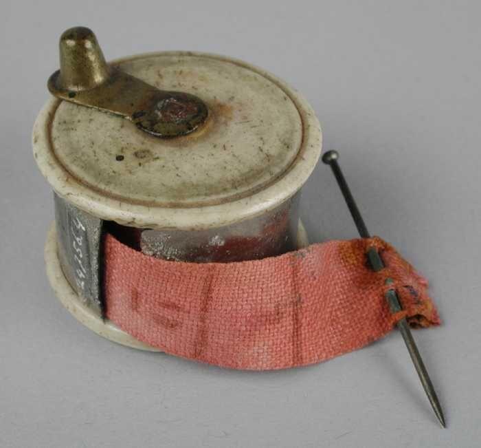 Klein rond doosje met opgerold meetlint, boven- en onderzijdevan ivoor, roze lint en messing…