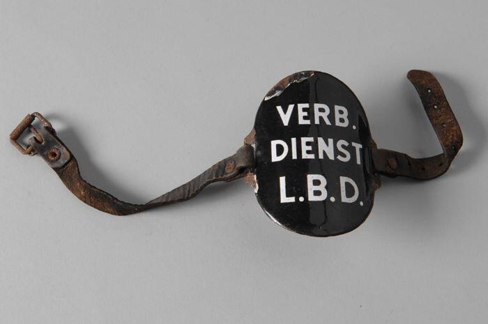 Ovaal zwart geëmailleerd armschildje met "VERB. DIENST L.B.D.", met leren riempjes aan weerszijde