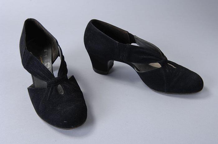 Zwarte suède hakschoenen, hoog gesloten op de wreef, vanaf de neus twee geplooide bandjes naar de…
