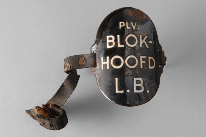 Ovaal zwart geëmailleerd armschildje met "PLV. BLOK-HOOFD L.B.", met (gescheurde) leren riempjes…