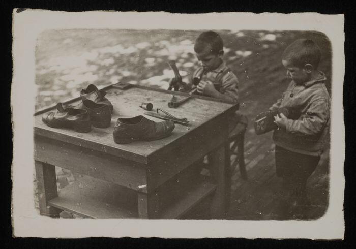 Zwart-wit foto van een buitenruimte met schoenmakerstafel waaraan twee kleine weesjongens van het…