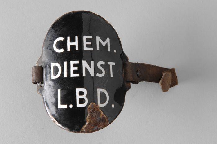 Ovaal zwart geëmailleerd armschildje met "CHEM. DIENST L.B.D.", met (gebroken) leren riempjes aan…