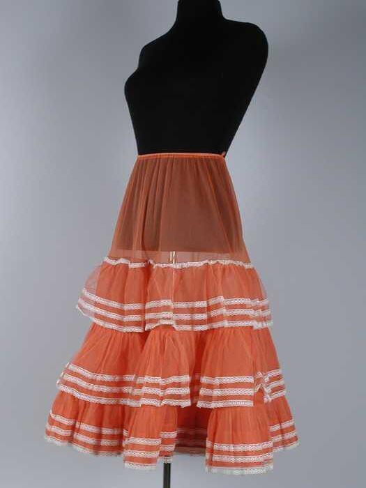 Wijde petticoat van lichtrood perlon, drie stroken tule met op elke baan drie witte kanten randjes,…
