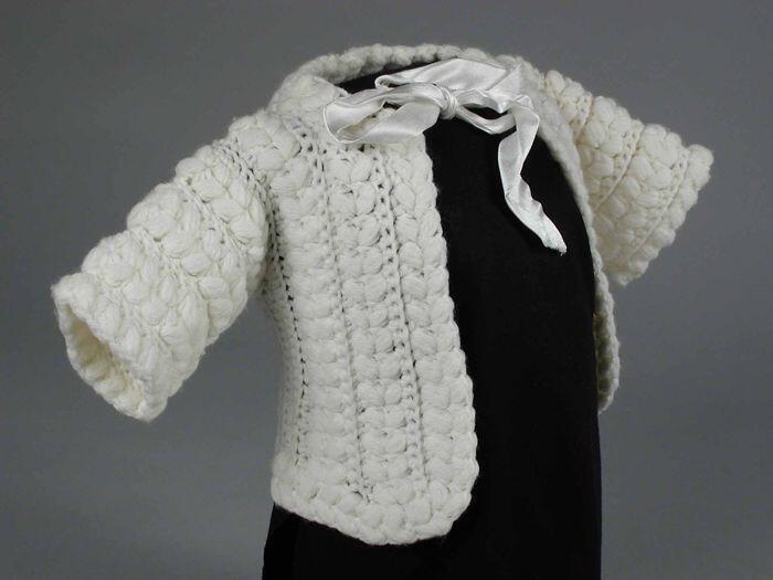 Babyvest gebreid van crèmekleurige wol in reliëfsteek met dikke noppen, wit satijnen strik bij de…