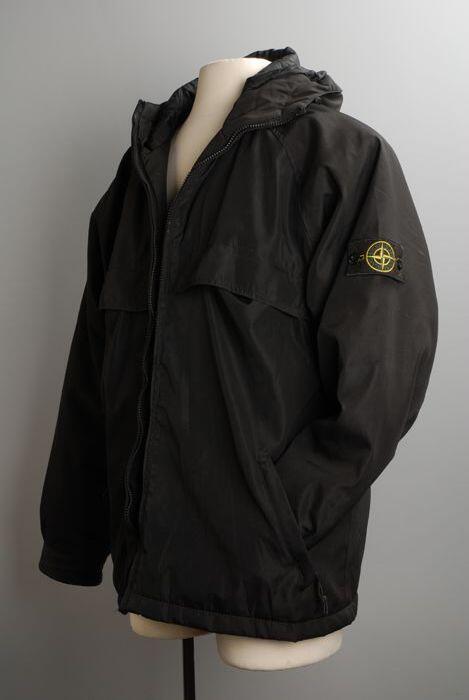 Lange zwarte jas met capuchon, merk Stone Island, met karakteristiek logo op de linkerarm