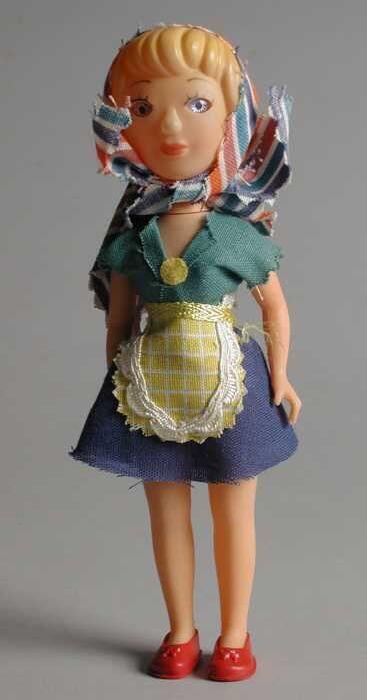 Kleine pop van roze gekleurd plastic, gekleed in blauwe rok, groen hesje en kleurige gestreepte…