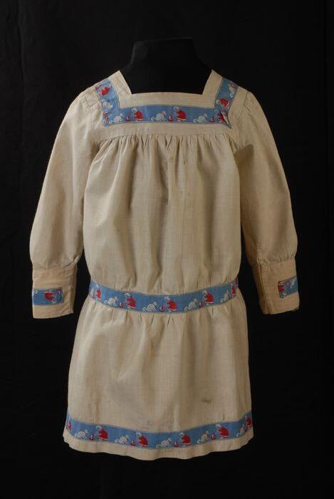 Kinderjurk, schort model van beige katoen met blauw sierband waarop een meisje dat een poesje melk…