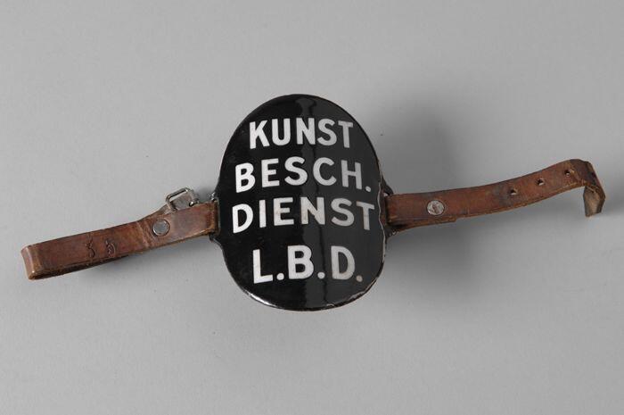 Ovaal zwart-geëmailleerd armschildje met "KUNST BESCH. DIENST L.B.D.", met leren riempjes aan…