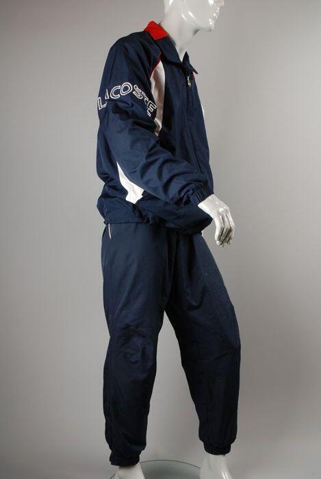 Blauwe trainingsjas van merk Lacoste, met merknaam in witte letters op de rechterarm en een klein…