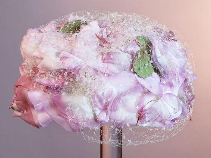 Ronde toque bedekt met roze en wit kunstzijde bloemen, waarover wit voile, hoedenspeld van metaal…