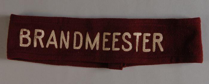 Hoedband van donkerrood laken met opdruk in wit, "BRANDMEESTER"
