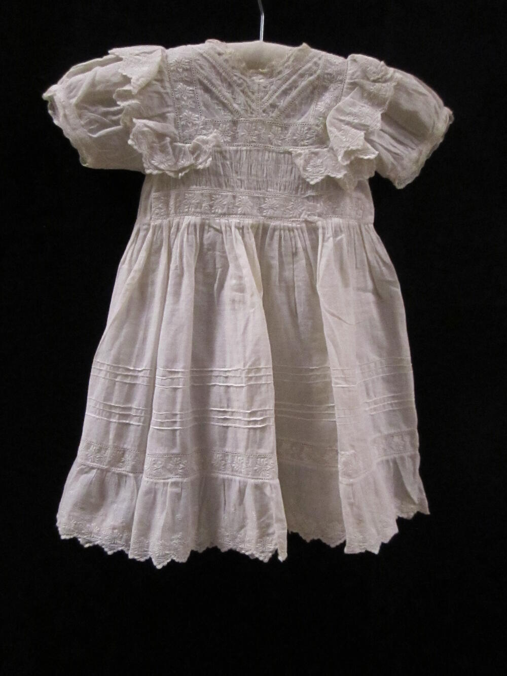 Jurk (kind) van witte broderie, kantrandjes langs hals, dubbele geschulpte volants broderie op de…