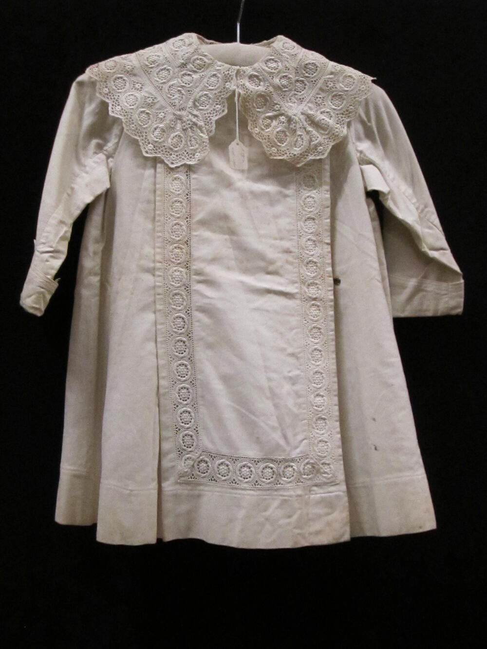 Jas (kind) van wit keperkatoen, geruwd aan de binnenzijde. Tussenzetsels van machinale broderie