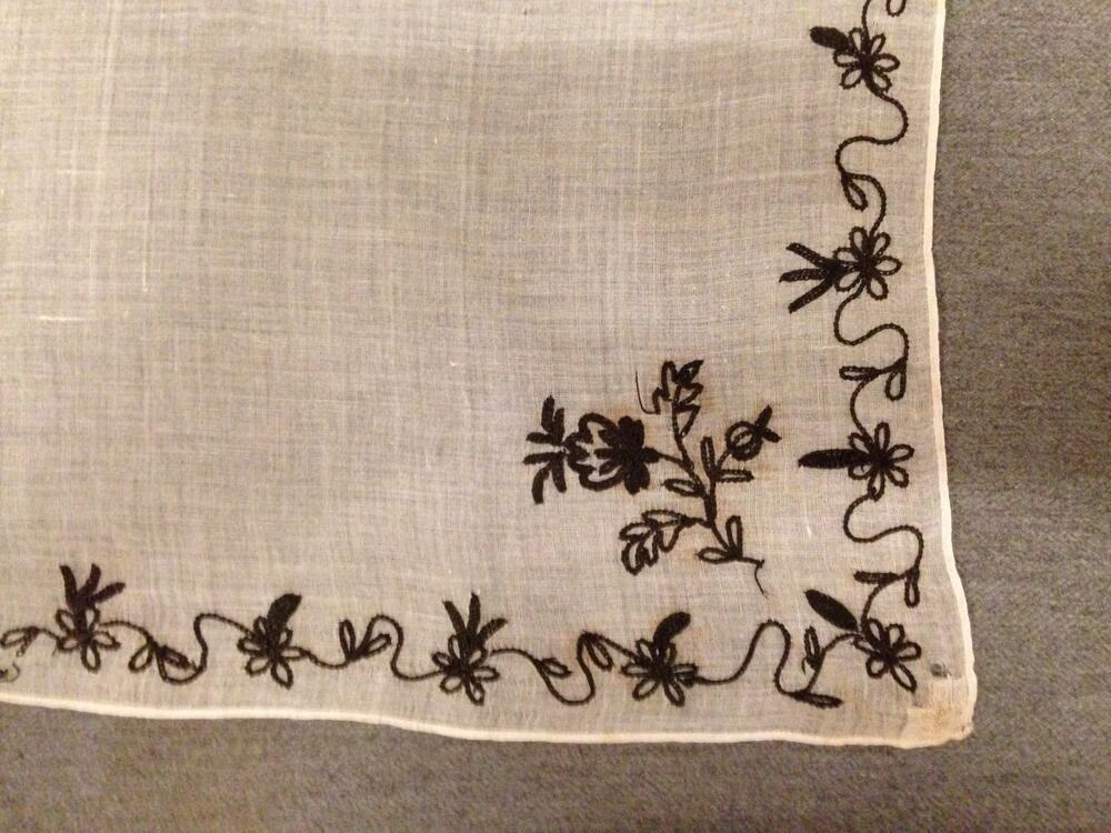 Tipdoekje, wit neteldoek getamboereerd met naief bloemrandje en hoekmotief in zwarte zijde. Rouw?