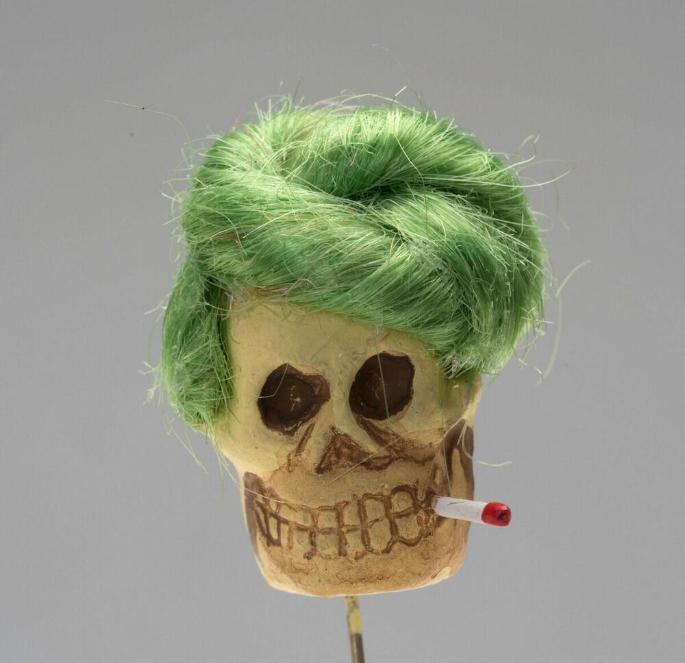 Kledingspeld in de vorm van een doodshoofd met groene pruik en sigaret, vervaardigd voor Allerzielen
