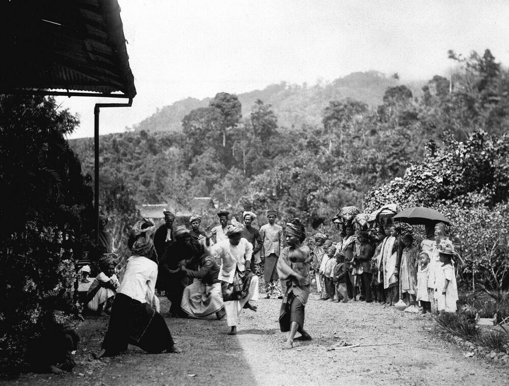 Een groep feestelijk geklede Indonesische mannen, vrouwen en kinderen bekijken een door gongs…