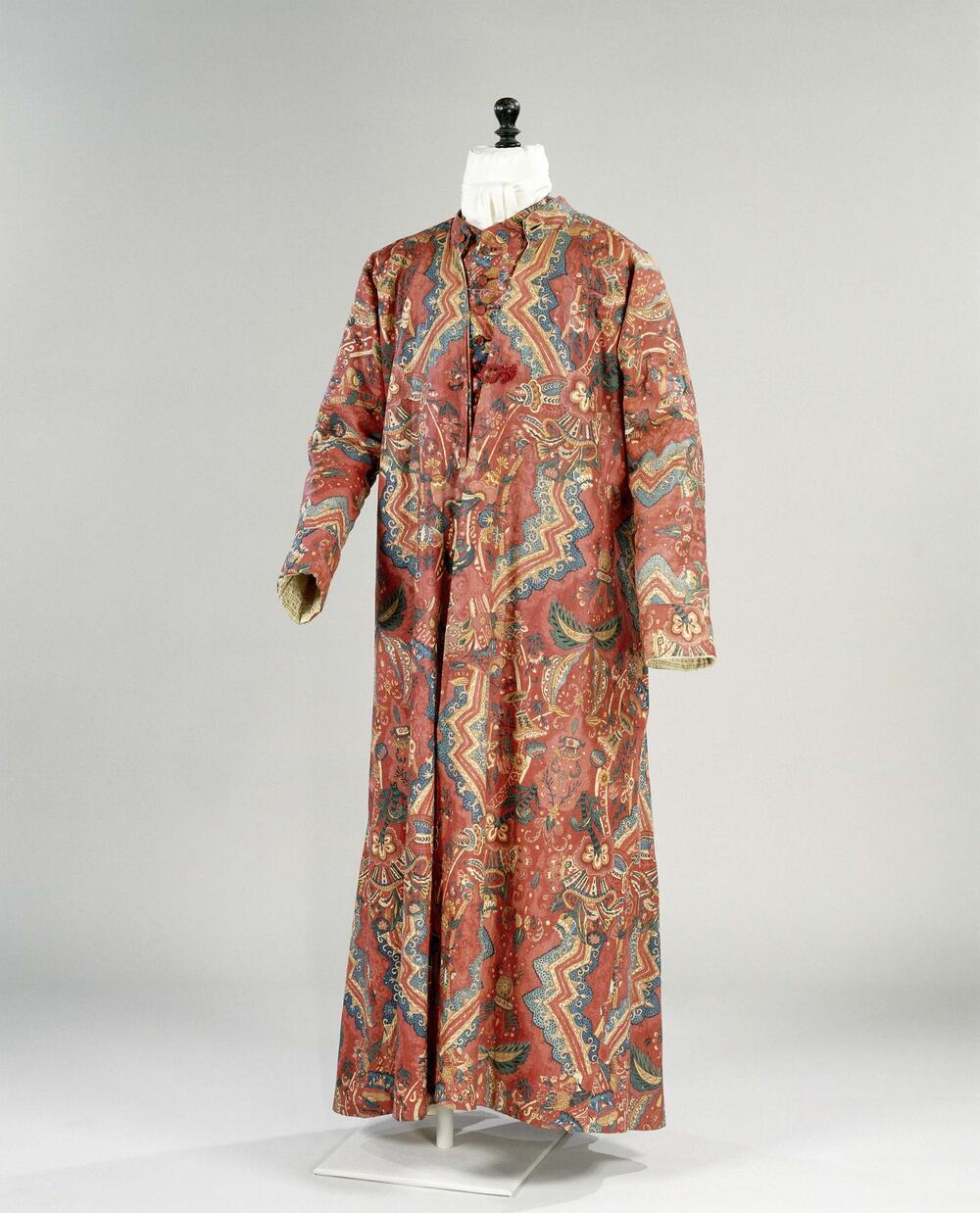Lang mouwloos vest van sits met een rood fond, versierd met gekleurde zigzagranden, omgeven door…