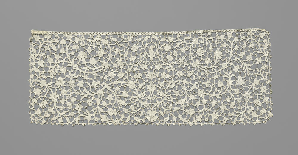 Slip van naaldkant met een symmetrisch patroon van dunne omkrullende ranken met veelsoortige bloemen