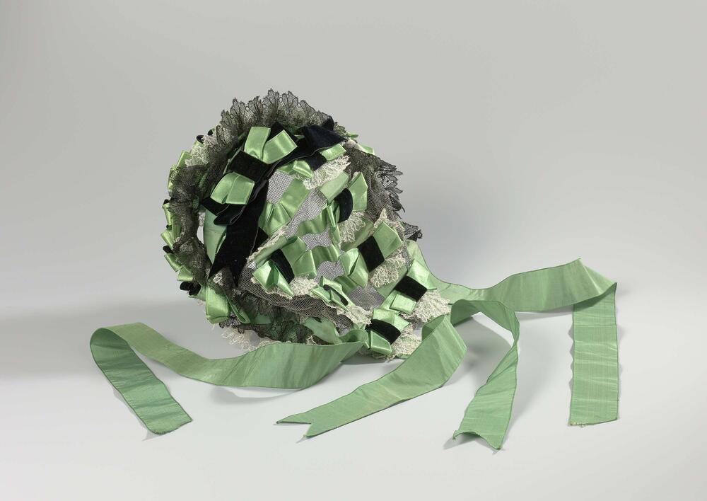 Bonnet-coiffure van groen satijn, zijden lint, bandfluweel en kant op een zwart tulen ondergrond…