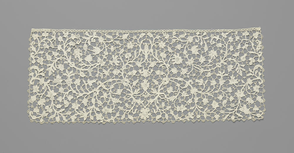 Slip van naaldkant met een symmetrisch patroon van dunne omkrullende ranken met veelsoortige bloemen