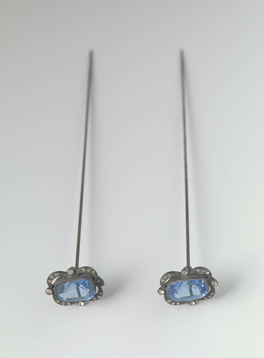 Hoedenspeld van staal met een langwerpig ornament van facet geslepen blauw glas gevat in een stalen…