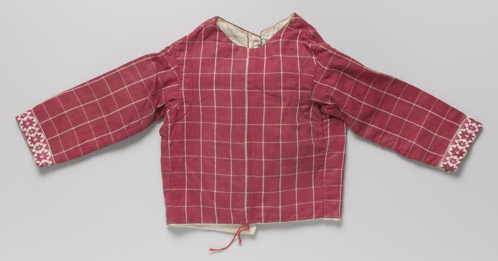 Rood linnen (?) jasje met wit ruitpatroon, langs de manchetten van de lange mouwen versierd met…