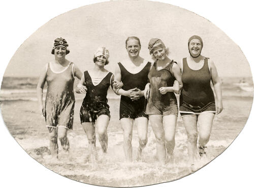 Badpakken. Een man tussen vier vrouwen, in badpak, rennend door de branding.Plaats en land onbekend. Badnummer 1923