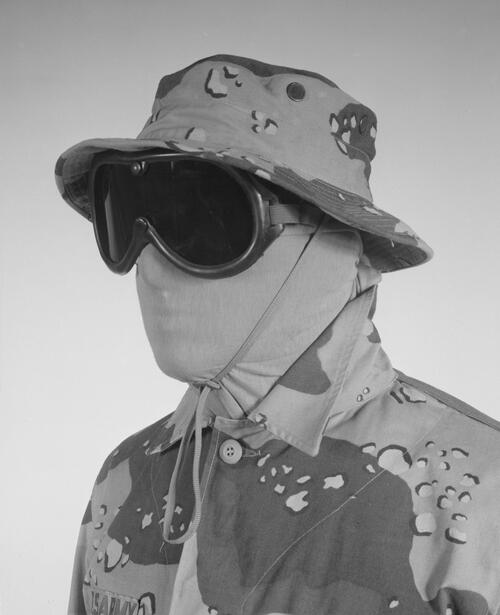 Zwart wit foto van man met camouflage leger outfit met vissershoed camouflage en groot gasmakerbril op en gezicht helemaal bedenkt met stof. Hij is geheel ingepakt